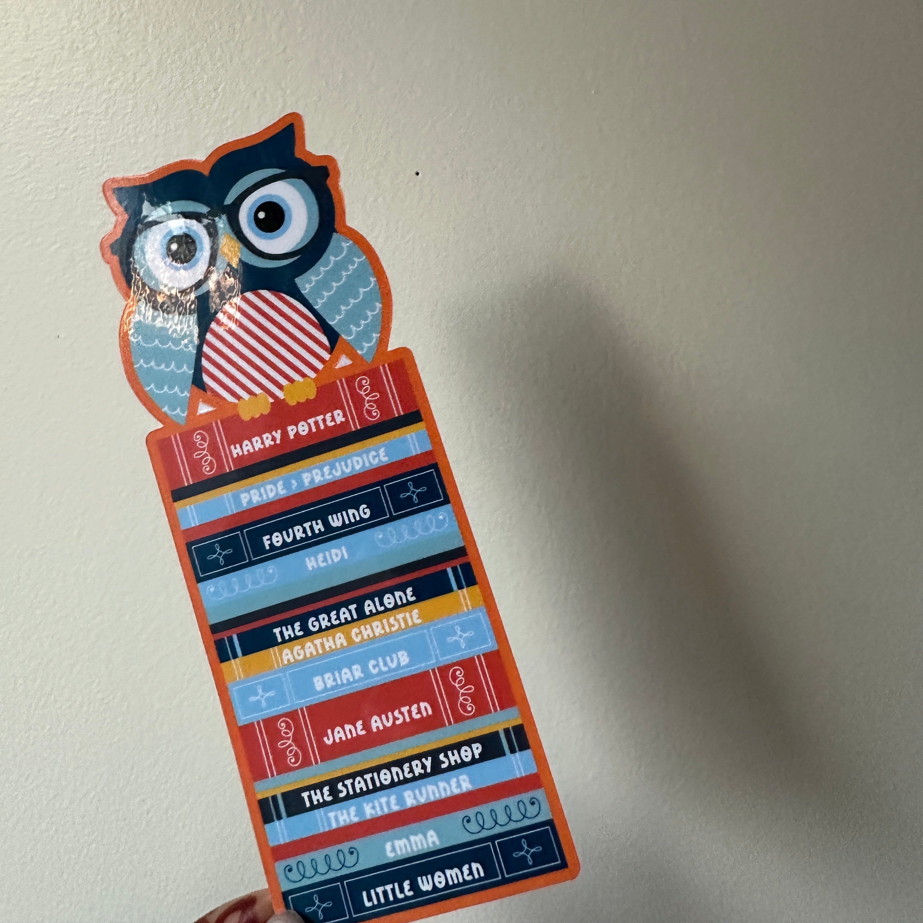 "Die-cut paper bookmark featuring an owl wearing glasses perched on a colorful stack of book spines with titles like Harry Potter, Pride and Prejudice, and Fourth Wing"