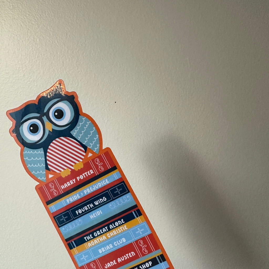 "Die-cut paper bookmark featuring an owl wearing glasses perched on a colorful stack of book spines with titles like Harry Potter, Pride and Prejudice, and Fourth Wing"