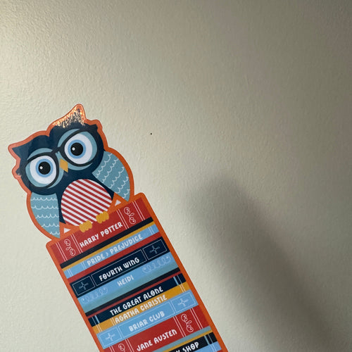 "Die-cut paper bookmark featuring an owl wearing glasses perched on a colorful stack of book spines with titles like Harry Potter, Pride and Prejudice, and Fourth Wing"