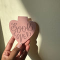 "Blush pink heart-shaped corner bookmark with hand-lettered 'Book Lover' text, designed to slip over a page corner"